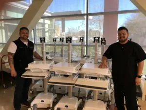 Two men standing beside medical carts with mounted tablet displays in a modern bright facility with large windows overlooking trees.
