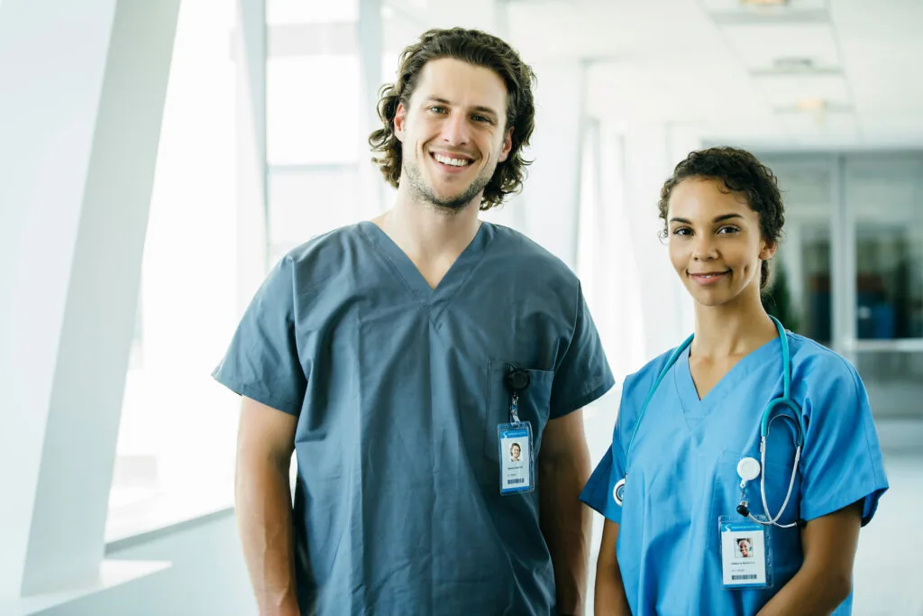 Two healthcare workers in scrubs and ID badges standing in a modern hospital corridor