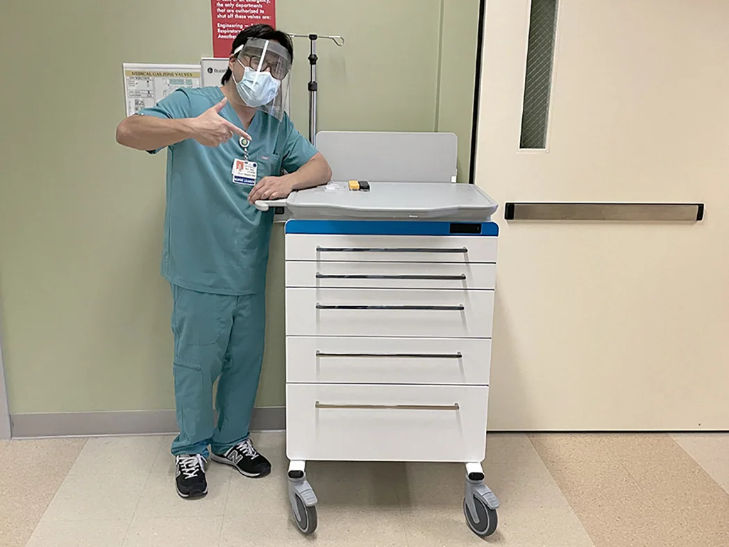 Healthcare worker in teal scrubs, face shield, and mask standing beside medical supply cart with drawers in hospital room